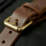 Brown leather backpack strap with a gold buckle on a dark background
