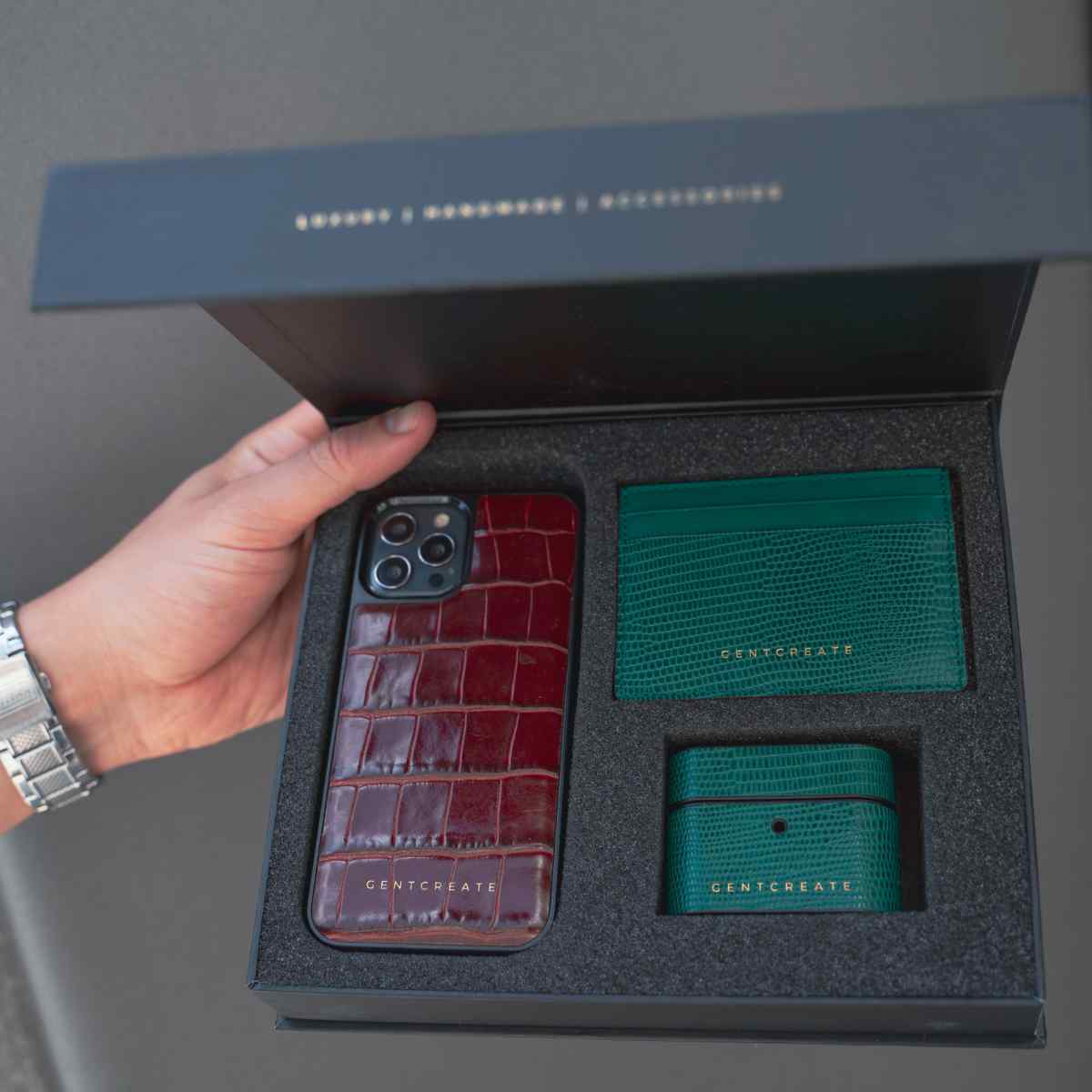 A man is holding a box from Gentcreate with luxury leather products inside, including an iPhone Leather Burgundy Case, a Green Cardholder and a Green AirPods case by Gentcreate