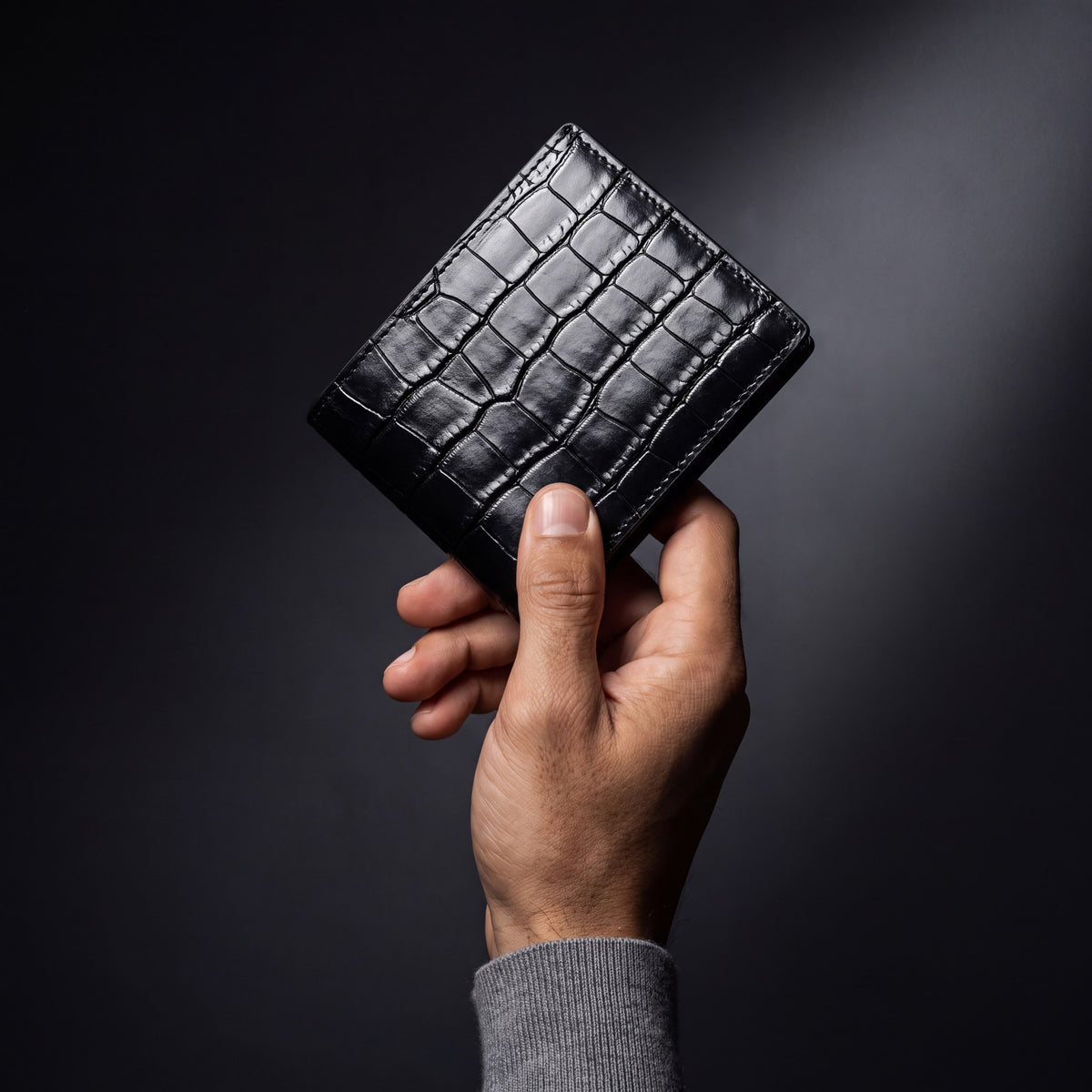 Man's hand holding a closed black crocodile-embossed leather bifold wallet by GENTCREATE in a dark studio