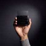 Man's hand holding a black crocodile-embossed leather card holder by GENTCREATE with a card in a dark studio