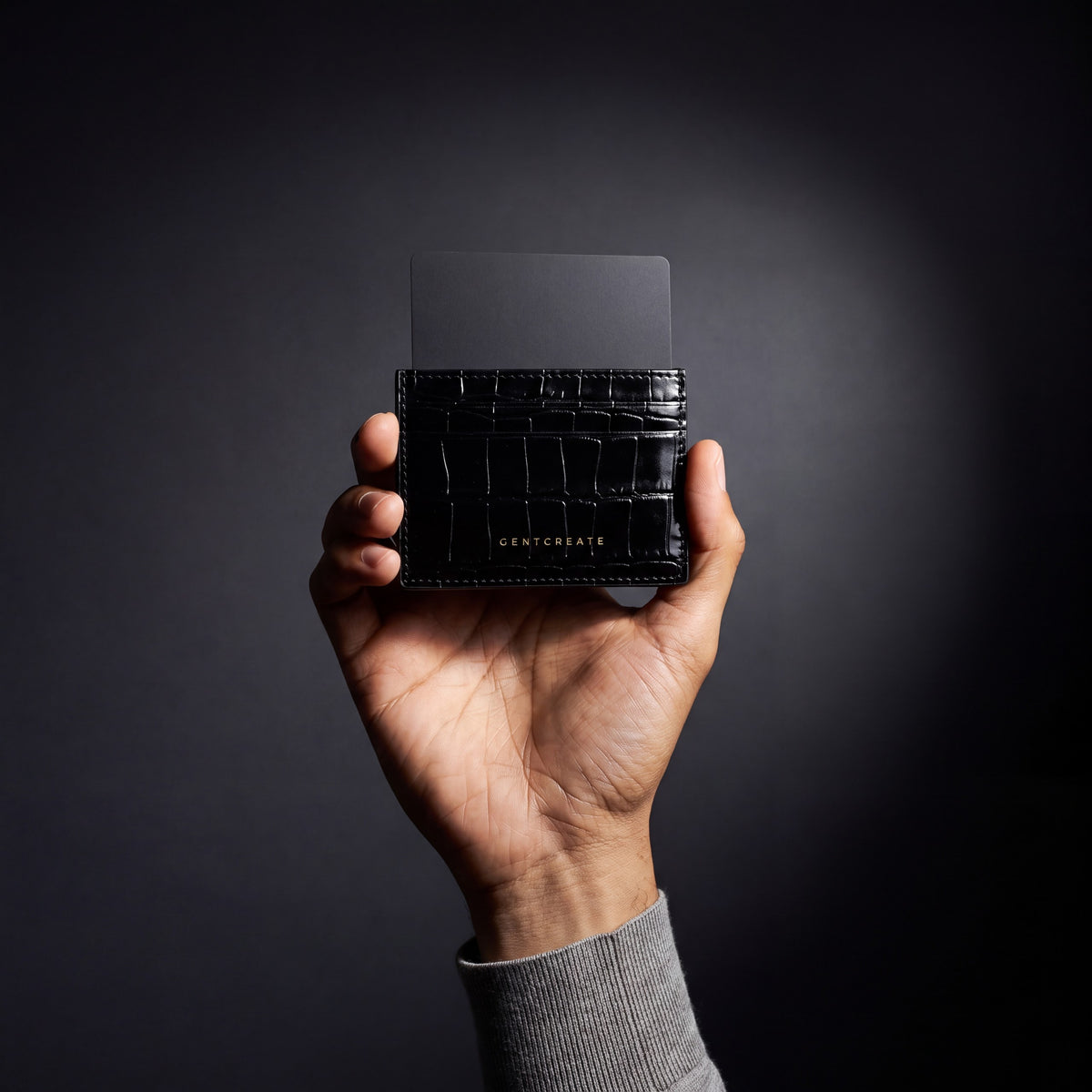 Man's hand holding a black crocodile-embossed leather card holder by GENTCREATE with a card in a dark studio