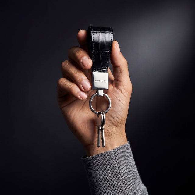 Man's hand holding up a black crocodile-embossed leather keychain by GENTCREATE with keys in a dark studio