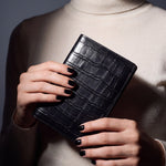Close-up of hands holding a black crocodile-embossed leather passport holder by GENTCREATE
