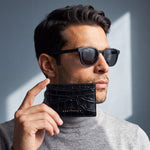 Man in sunglasses holding a black glossy crocodile-embossed leather card holder by GENTCREATE against his jaw in window light