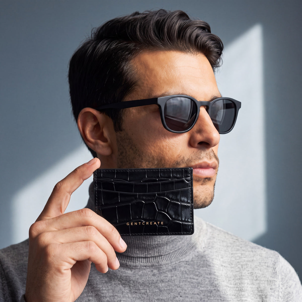 Man in sunglasses holding a black glossy crocodile-embossed leather card holder by GENTCREATE against his jaw in window light