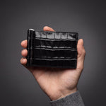 Man's hand holding a black glossy crocodile-embossed leather money clip wallet by GENTCREATE in a dark studio