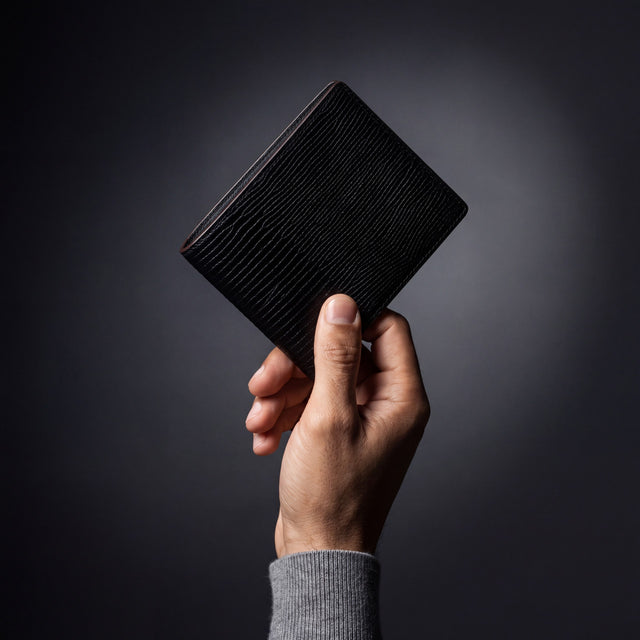 Man's hand holding a black lizard-embossed leather bifold wallet by GENTCREATE in a dark studio