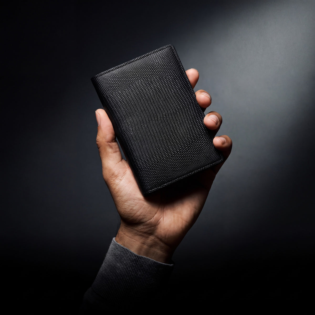 Man's hand holding a black lizard-embossed leather passport holder by GENTCREATE in a dark studio