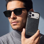 رجل يرتدي نظارة شمسية يمسك جرابًا أسود لهاتف iPhone 18 Pro Max من جلد عجل بنقش موجي بارز من GENTCREATE بجانب وجهه تحت ضوء النافذة