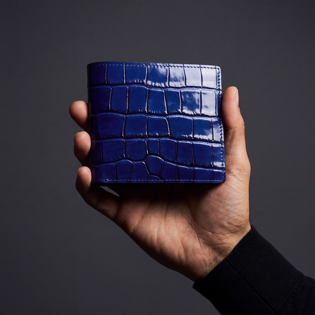 Main d'homme tenant un portefeuille GENTCREATE à deux volets en cuir bleu brillant embossé façon crocodile, en studio sombre