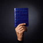 יד גברית אוחזת בנרתיק דרכון כחול מעור מבריק מוטבע בדוגמת תנין מבית GENTCREATE, בסטודיו חשוך