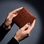 Close-up of hands holding a brown crocodile-embossed leather bifold wallet by GENTCREATE