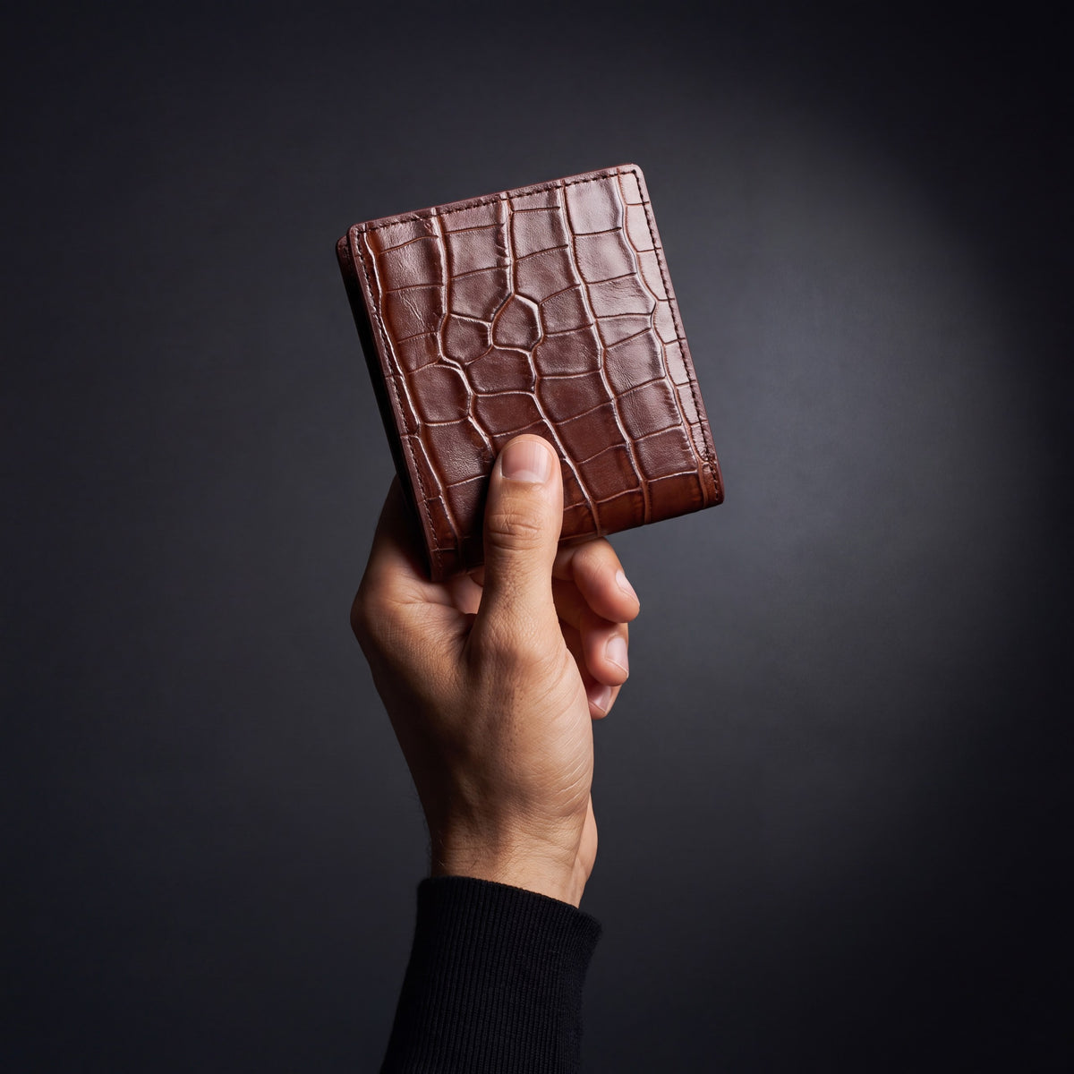 Man's hand holding a closed brown crocodile-embossed leather bifold wallet by GENTCREATE in a dark studio