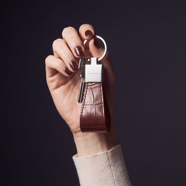 Woman's hand holding up a brown crocodile-embossed leather keychain by GENTCREATE by its split ring in a dark studio