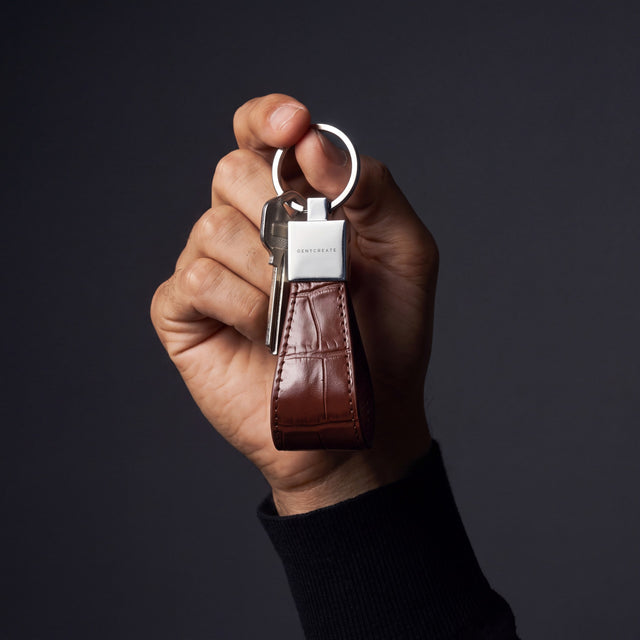 Man's hand holding up a brown crocodile-embossed leather keychain by GENTCREATE with keys in a dark studio