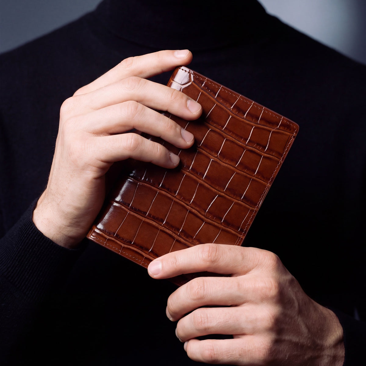 Close-up of hands holding a brown crocodile-embossed leather passport holder by GENTCREATE