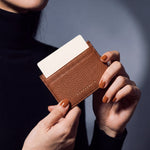 Close-up of hands holding a brown pebble leather card holder by GENTCREATE with a card in the top slot