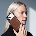 Kvinde holder et brunt iPhone 18 Pro Max-cover i narvet kalveskind fra GENTCREATE ind mod kinden i dagslys fra et vindue