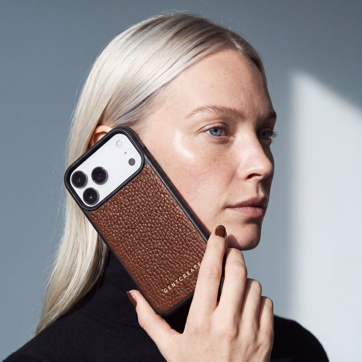 Kvinde holder et brunt iPhone 18 Pro Max-cover i narvet kalveskind fra GENTCREATE ind mod kinden i dagslys fra et vindue