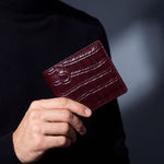 Close-up of hands holding a burgundy crocodile-embossed leather bifold wallet by GENTCREATE
