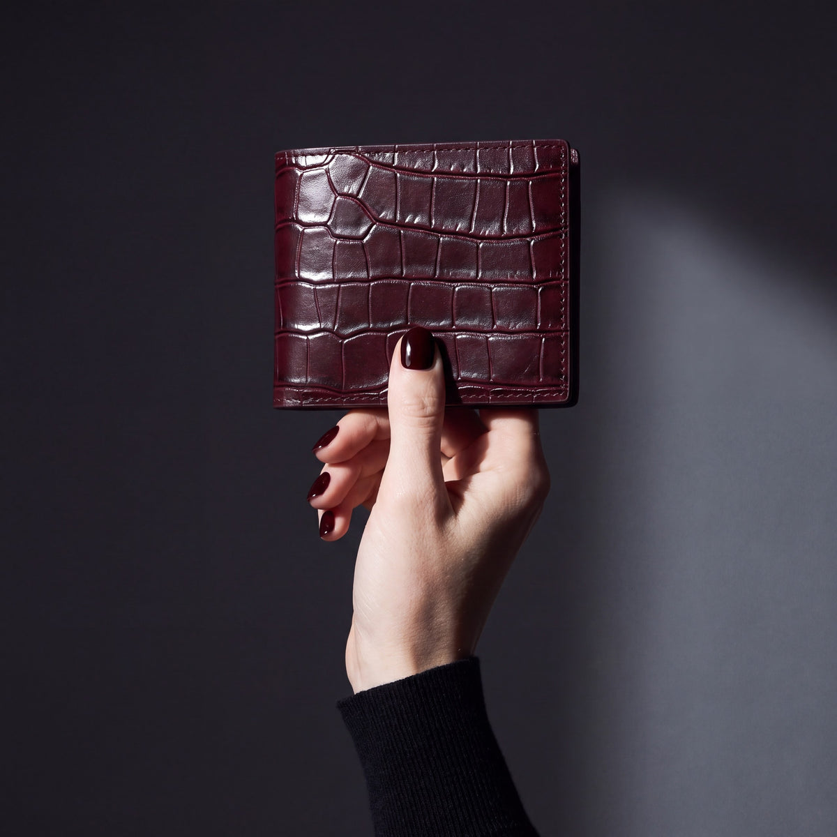 Woman's hand holding a closed burgundy crocodile-embossed leather bifold wallet by GENTCREATE in a dark studio