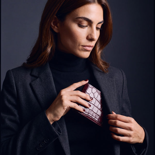 Woman sliding a burgundy crocodile-embossed leather bifold wallet by GENTCREATE into a blazer pocket