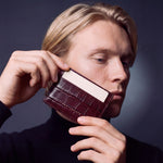 Man holding a burgundy crocodile-embossed leather card holder by GENTCREATE by his face