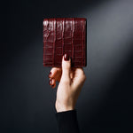 Main de femme tenant un portefeuille GENTCREATE à deux volets en cuir bordeaux brillant embossé façon crocodile, en studio sombre
