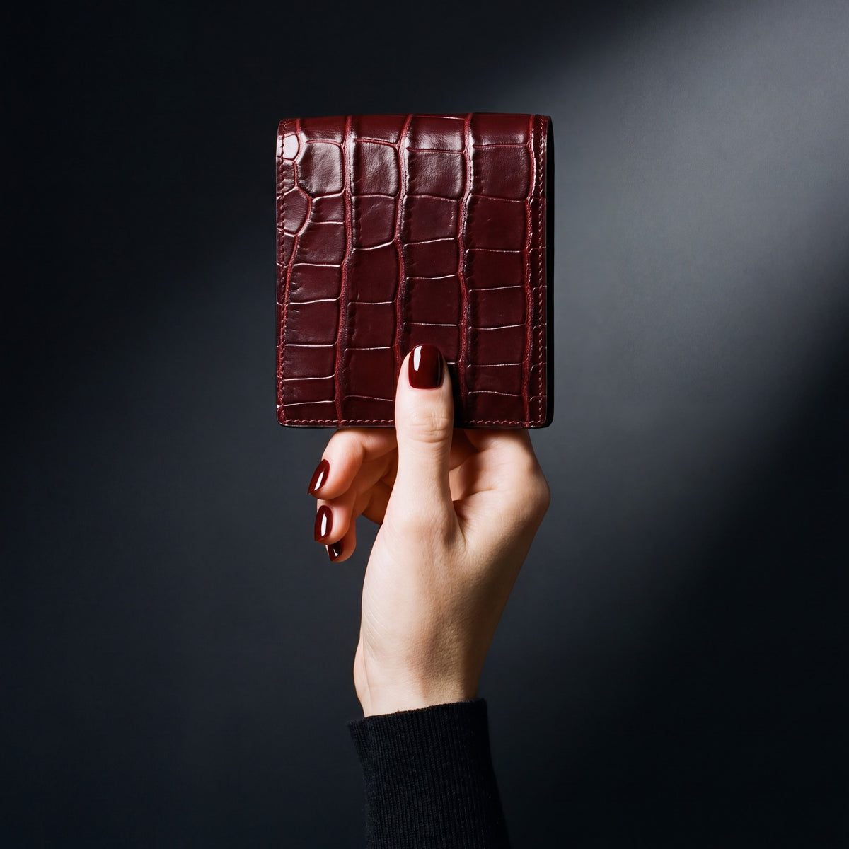 Main de femme tenant un portefeuille GENTCREATE à deux volets en cuir bordeaux brillant embossé façon crocodile, en studio sombre