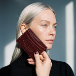 Woman holding a burgundy glossy crocodile-embossed leather card holder by GENTCREATE to her cheek in window light