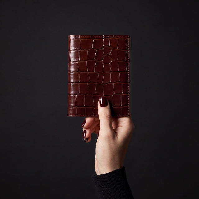 Mão feminina a segurar um porta-passaporte da GENTCREATE em pele bordeaux brilhante gravada efeito croco, em estúdio escuro