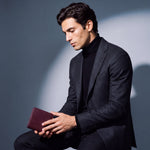 Man seated, holding a burgundy lizard-embossed leather bifold wallet by GENTCREATE