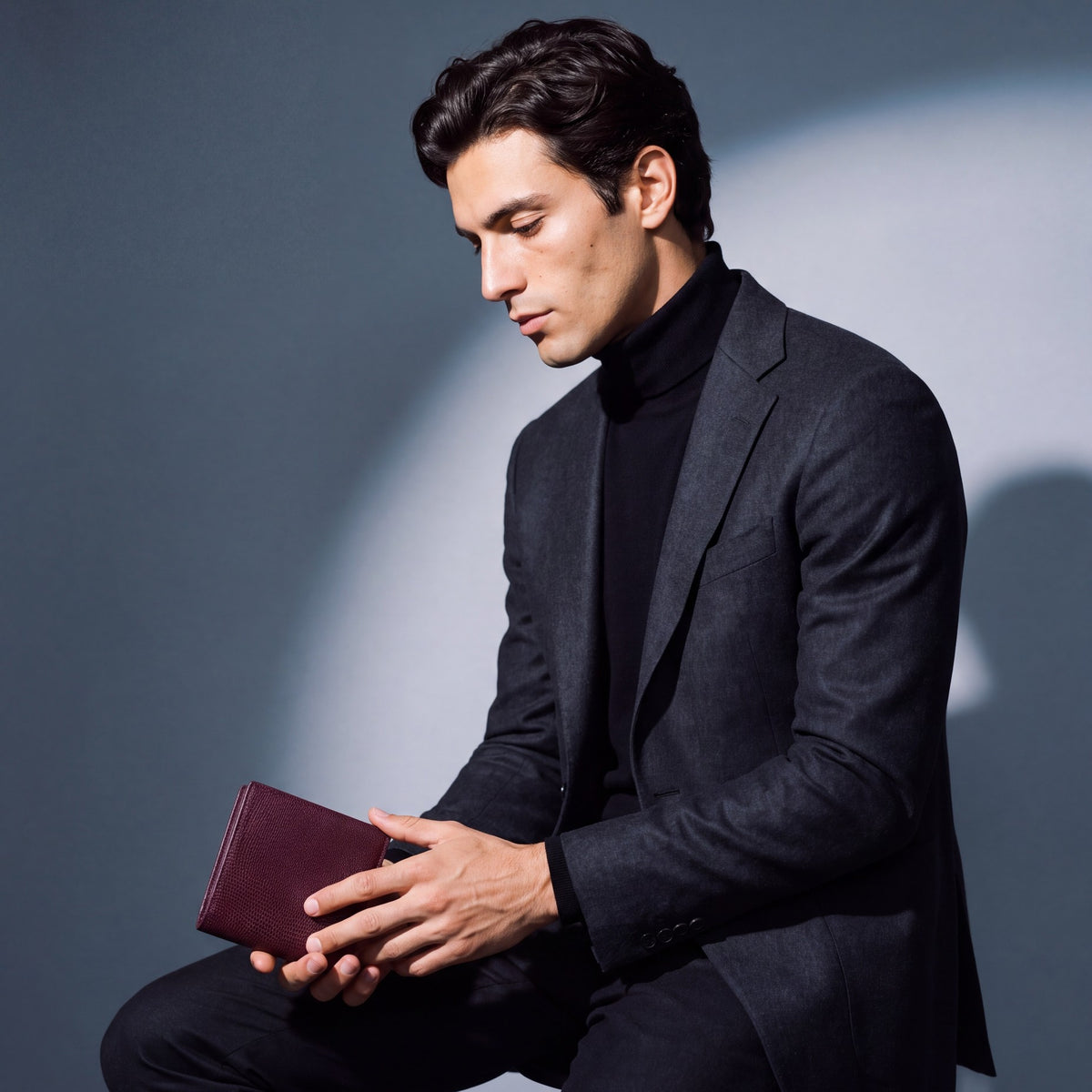Man seated, holding a burgundy lizard-embossed leather bifold wallet by GENTCREATE