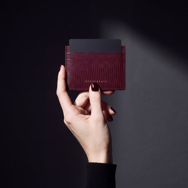 Woman's hand holding a burgundy lizard-embossed leather card holder by GENTCREATE with a card in a dark studio