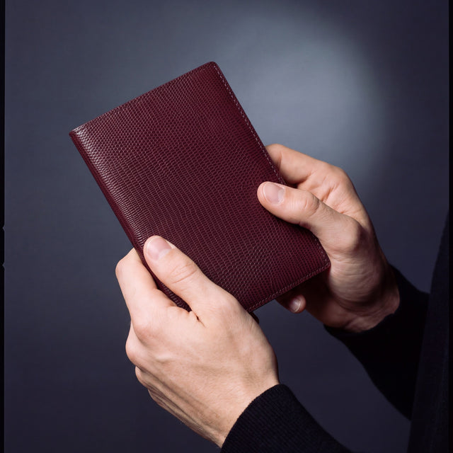 Close-up of hands holding a burgundy lizard leather passport holder by GENTCREATE