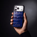 Man's hand holding a blue glossy crocodile-embossed calfskin iPhone 18 Pro Max case by GENTCREATE in a dark studio
