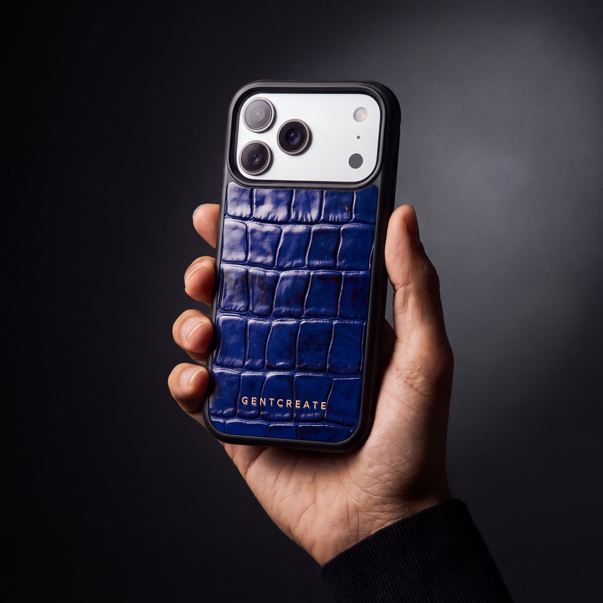 Man's hand holding a blue glossy crocodile-embossed calfskin iPhone 18 Pro Max case by GENTCREATE in a dark studio
