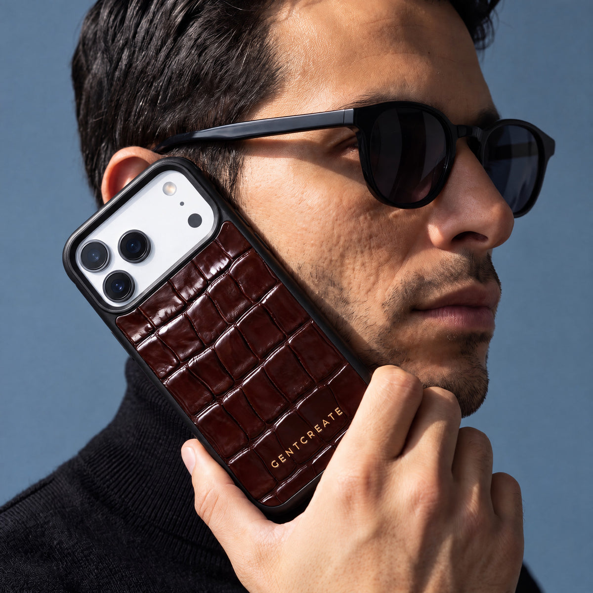 Homme à lunettes de soleil tenant contre sa joue une coque iPhone 18 Pro Max GENTCREATE en veau bordeaux brillant embossé façon crocodile