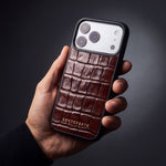 Man's hand holding a burgundy glossy crocodile-embossed calfskin iPhone 18 Pro Max case by GENTCREATE in a dark studio