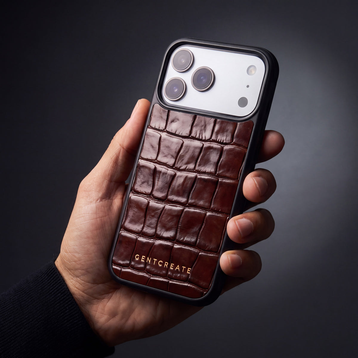 Man's hand holding a burgundy glossy crocodile-embossed calfskin iPhone 18 Pro Max case by GENTCREATE in a dark studio