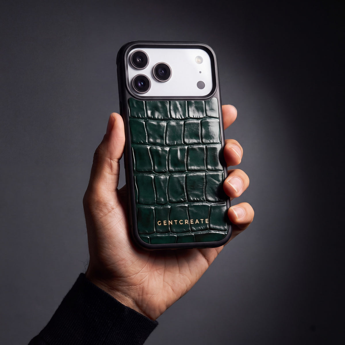 Man's hand holding a green glossy crocodile-embossed calfskin iPhone 18 Pro Max case by GENTCREATE in a dark studio