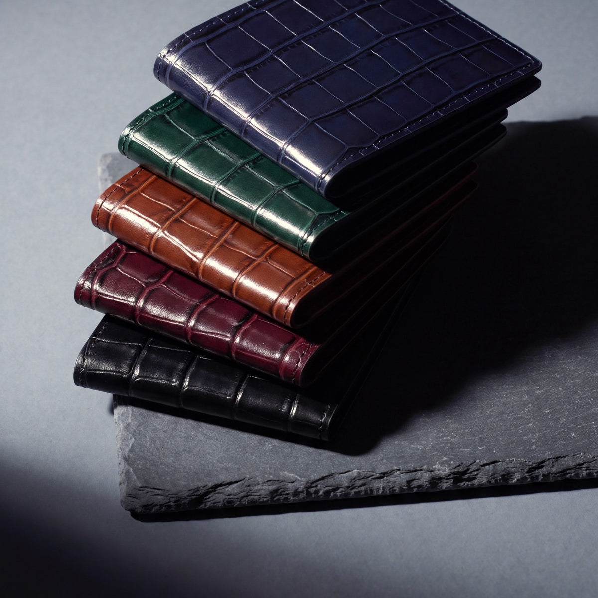 Five crocodile-embossed GENTCREATE wallets stacked on slate: navy, green, brown, burgundy and black