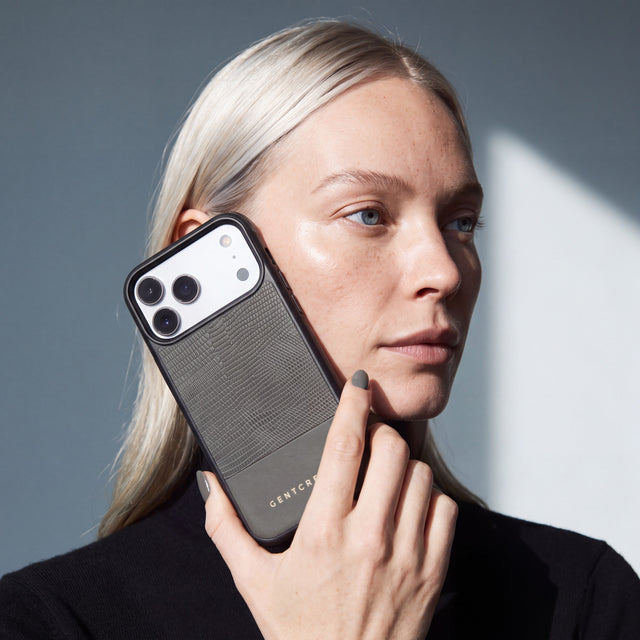 امرأة تمسك جرابًا رماديًا لهاتف iPhone 18 Pro Max من جلد عجل بنقش السحلية البارز من GENTCREATE مسندًا إلى خدها تحت ضوء النافذة