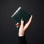 Woman's hand holding a closed green crocodile-embossed leather bifold wallet by GENTCREATE in a dark studio
