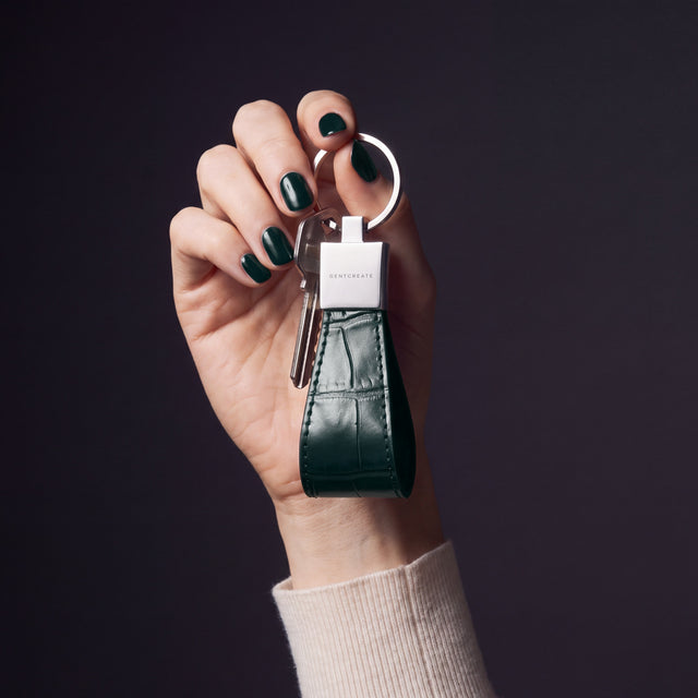 Woman's hand holding up a green crocodile-embossed leather keychain by GENTCREATE by its split ring in a dark studio