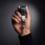 يد رجل ترفع ميدالية مفاتيح خضراء من جلد بنقش التمساح البارز من GENTCREATE مع مفاتيح في استوديو مظلم