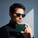 Man in sunglasses holding a green glossy crocodile-embossed leather card holder by GENTCREATE below his chin in window light