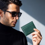Homme à lunettes de soleil tenant à hauteur de poitrine un porte-cartes de visite GENTCREATE en cuir vert embossé façon lézard, à la lumière d'une fenêtre