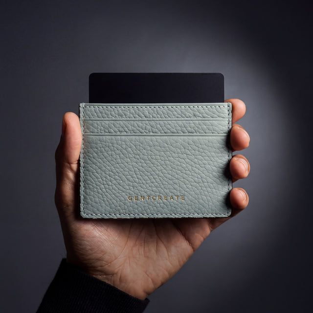 Main d'homme tenant un porte-cartes GENTCREATE en cuir grainé gris clair avec une carte, en studio sombre
