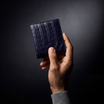 Man's hand holding a closed navy crocodile-embossed leather bifold wallet by GENTCREATE in a dark studio